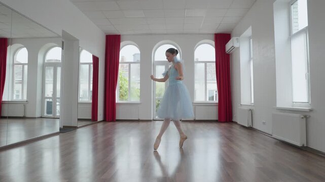 Young elegant ballerina in a blue tutu gracefully performing a classical dance routine on pointe shoes in a spacious, light-filled studio with large windows and a mirror wall
