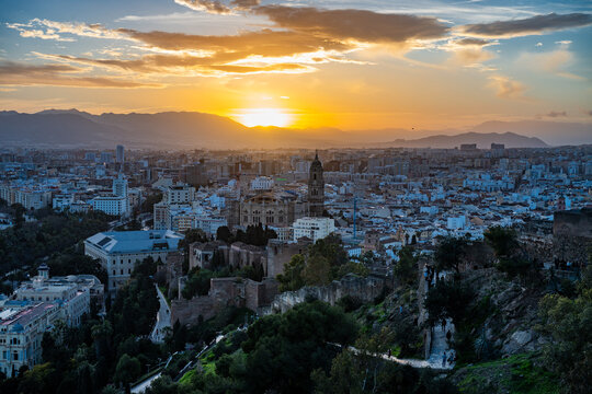 Wide panoramic view of Malaga city from a hilltop at sunset, showing the cathedral, historical buildings, and mountains under a warm, cloudy sky.