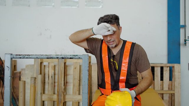 Exhausted male construction worker taking off yellow hard hat and wiping sweat from forehead while resting in an industrial warehouse. Tired factory laborer in safety vest on a hot work break.