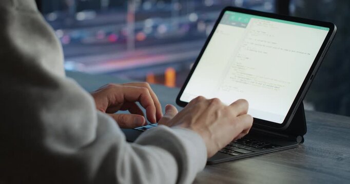 A man in a grey hoodie types code on a tablet keyboard at a wooden table with a blurred city street behind him.