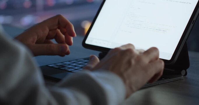 Close-up of hands typing on a tablet keyboard with lines of code on the bright screen and soft city bokeh behind.