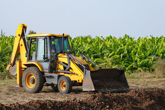 A yellow backhoe loader is digging and leveling soil in a rural agricultural field