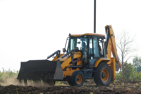 A yellow backhoe loader is digging and leveling soil in a rural agricultural field