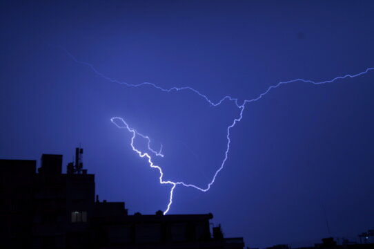 Brilliant forked lightning illuminates a deep blue night sky above black urban silhouettes. The image represents raw power, urgency, and a dramatic moment in nature
