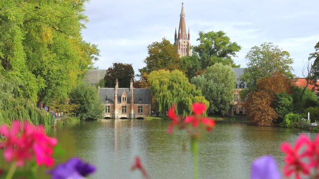 Bruges town in Belgium, Europe. Minnewater Lake Serenity: Beguinage and Church Spire Framed by Lush Greenery and Vibrant Flowers on a Calm Autumn Day