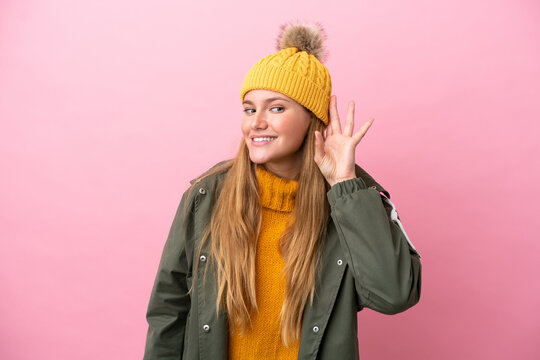 Young blonde woman wearing winter jacket isolated on pink background listening to something by putting hand on the ear