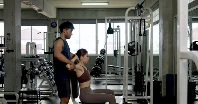 Young Asian couple exercising together in modern gym, man standing behind seated woman while guiding dumbbell curl form, partner training with support, motivation, healthy lifestyle teamwork.