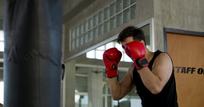 Adult Caucasian man strikes heavy bag with red boxing gloves in modern gym, alternating guard and punches. Male athlete builds power, endurance, coordination, confidence, active workout routine.