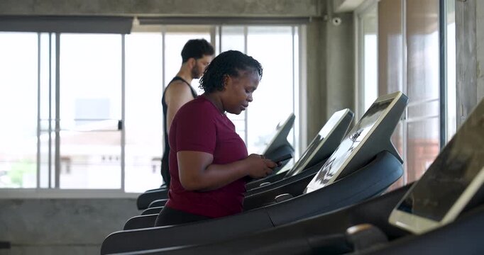 African American plus size woman jogging at steady pace on treadmill near bright window in gym. Curvy female athlete doing cardio exercise for endurance, wellness, confidence, healthy living.