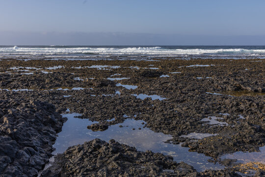 Expansive field of dark volcanic tide pools reflecting sky under distant waves