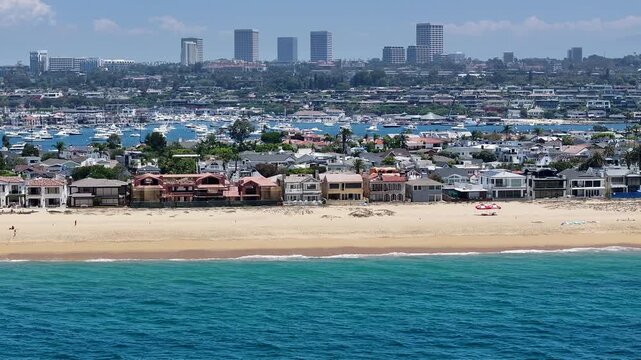 flying southbound along Balboa Beach located on Harbor Island, a small artificial island in Newport Beach, California, while massive waves hitting the wide sandy beach and people sunbathing