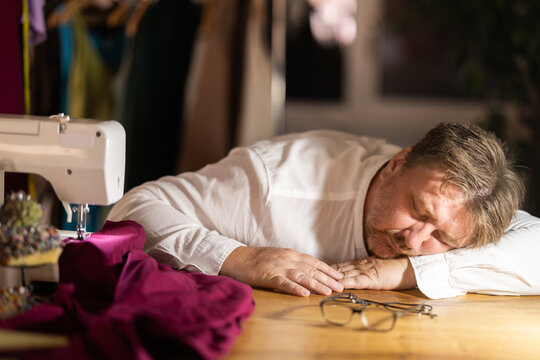 Overworked middle-aged male tailor sleeping on table next to sewing machine