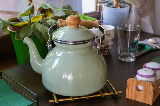 A mint green vintage-style enamel tea kettle with wooden handles sits on a trivet on a dark table, surrounded by potted houseplants, a drinking glass, and salt and pepper shakers