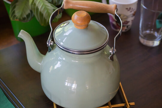 a mint green vintage-style enamel tea kettle with wooden handles resting on a trivet on a dark kitchen table, softly framed by a potted plant and drinking glasses