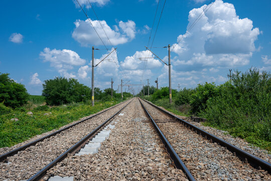 a symmetrical, eye-level perspective down the center of empty double railway tracks stretching toward a vanishing point on the horizon, flanked by green vegetation under a bright blue sky filled
