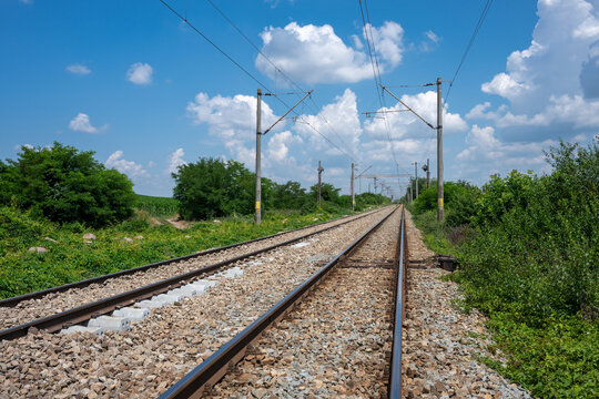 empty double railway tracks stretching toward the horizon, bordered by green vegetation and utility poles under a partly cloudy blue sky.