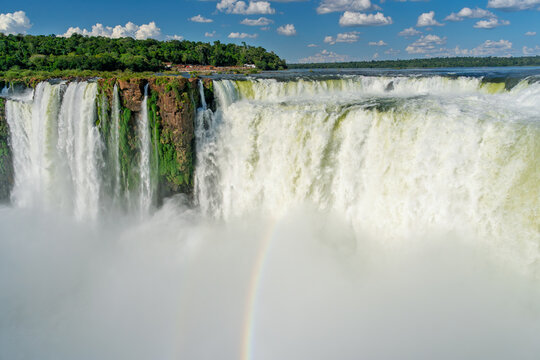 Scenic view of a massive waterfall in Brazil with a rainbow in the mist and lush green forest under a blue sky