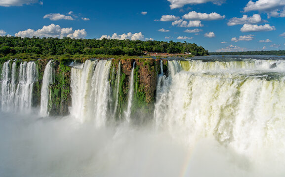 Panoramic view of a massive waterfall in the Brazilian rainforest with lush green vegetation and a rainbow under a blue sky