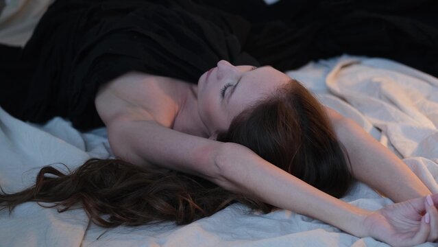 A portrait of a beautiful brunette with long hair, wearing a black tunic or chiton dress, in a studio. The beautiful, natural woman poses for the camera while lying on the floor.