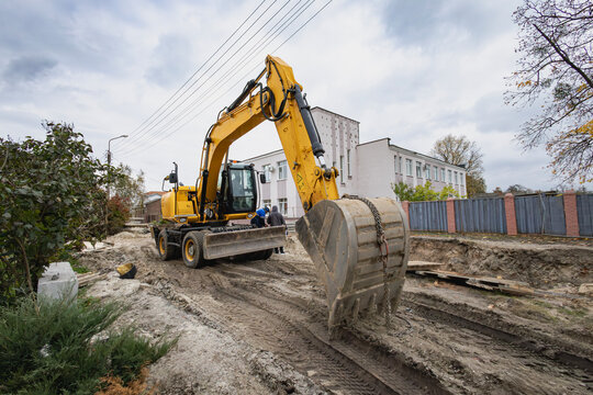 A yellow excavator works on a construction site digging into the ground. Nearby, there are trees, a building, and utility poles visible under cloudy skies