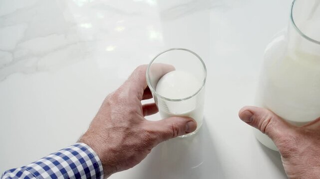 Man's hand pours fresh milk from a glass bottle into a clear glass on a white marble table. Symbolizes dairy nutrition, healthy breakfast, and organic farming. Ideal for food and wellness content