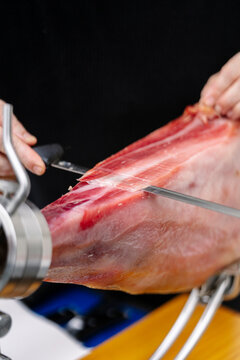 Close-up of hands slicing cured ham with knife, natural photo style on dark background, concept of culinary preparation