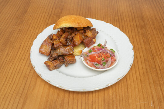 Bread roll with fried pork belly chunks and tomato onion salsa on a white plate over wood.