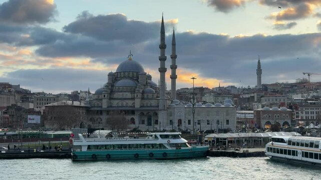 Establishing water-level shot of the monumental New Mosque and its iconic minarets during a vibrant sunset at the historic Eminonu waterfront in Istanbul, Turkey.
