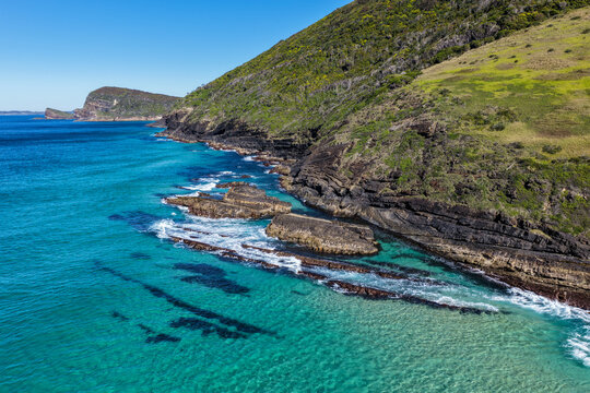 Aerial view of the clear turquoise waters surrounding Blueys Head at Blueys Beach, with the rocky headland, pristine sandy shoreline and vibrant coastal waters along the Mid North Coast of NSW.