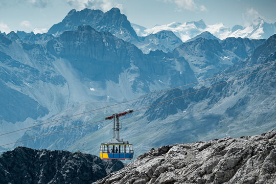 A colorful cable car travels above rugged alpine terrain, surrounded by dramatic rocky peaks in the Swiss alps, snow-covered mountains, and glaciers in a breathtaking high-altitude landscape.