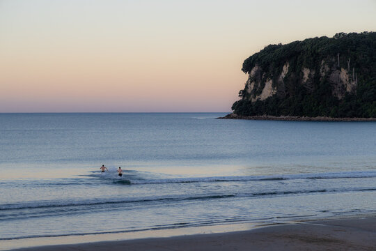 Three friends walk into the ocean at sunset on Whangamata Beach, Coromandel Peninsula, New Zealand. The iconic Clark island is visible in the background.
