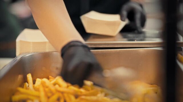 A cook in an apron and gloves places French fries into a paper container in a bright kitchen. Concept for preparing and packing takeout orders in a fast food establishment