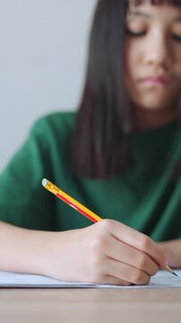Vertical video close up and selective focus showing Asian kid girl concentrating as she writes in her notebook with a pencil. Bright daylight fills the room while she is doing her homework.