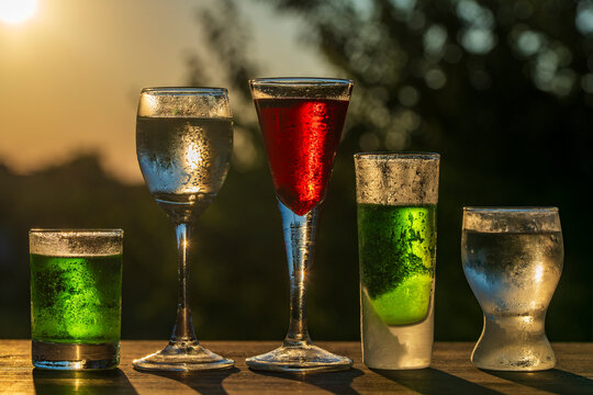 Set of various alcoholic drinks, liqueurs, and shots in mismatched glasses on wooden table outdoors, closeup. Bright green, red, and clear beverages backlaced by warm evening sunset light