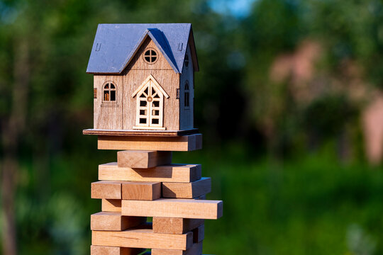 Toy wooden house on top of unstable tower made of jenga blocks outdoors. Metaphor for real estate risk, property market instability, mortgage crisis, finance, investment, or building foundation