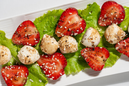 Gourmet summer salad with ripe strawberries mozzarella cheese balls green lettuce balsamic glaze black pepper and sesame seeds on white plate, closeup. Healthy organic food and fine dining concept