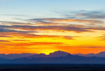 Fototapeta premium landscape background of sunset sky featuring layers of orange and yellow clouds above a dark mountain range silhouette in a peaceful evening.