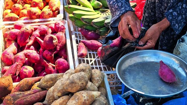 A variety of fresh sweet potatoes and colorful bananas at a local open-air market, with vendors preparing to buy. Shopping for traditional foods. 4K