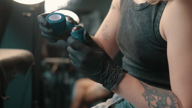 Tattoo artist wearing black gloves wrapping blue grip tape around a tattoo machine before a professional tattooing session in a studio.