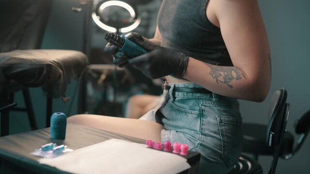 Tattoo artist wearing black gloves wrapping blue grip tape around a tattoo machine before a professional tattooing session in a studio.