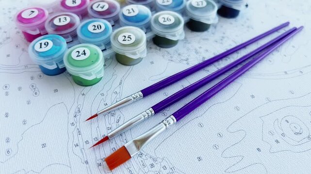 The camera smoothly moves away from the canvas with numbers, showing purple brushes and jars of acrylic paint. The concept of creativity, hobbies, art therapy, relaxation, DIY paintings, development