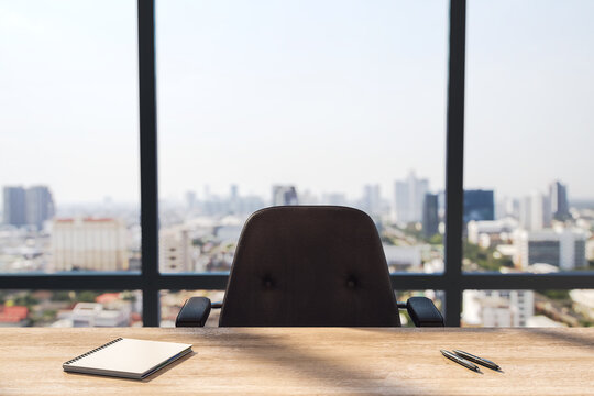 Wooden desk with spiral notepad and pens, dark leather chair facing panoramic window with city view in empty office workplace. 3D Rendering