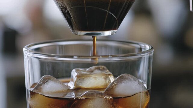 Dark espresso drips from a pour over coffee dripper into a clear glass filled with ice.