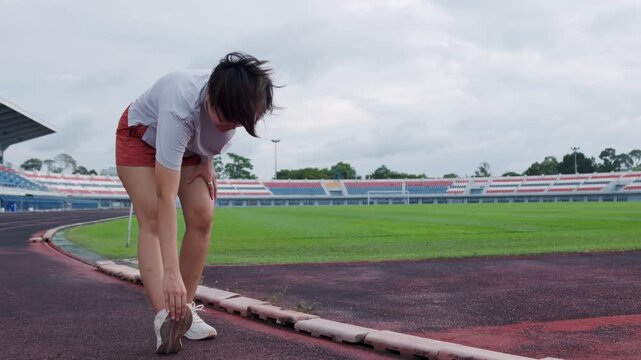 Authentic shot of an active Asian woman stretching on a running track beside a green soccer field. She bends down, providing excellent copy space for your health and fitness project backgrounds.