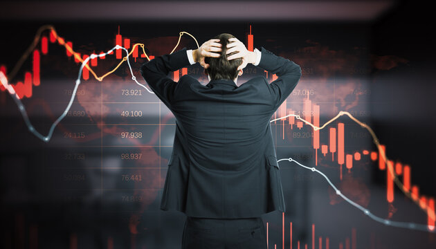 Stressed businessman holding head in hands facing screen with red falling candlestick charts and numbers, stock market crash and trader panic concept.