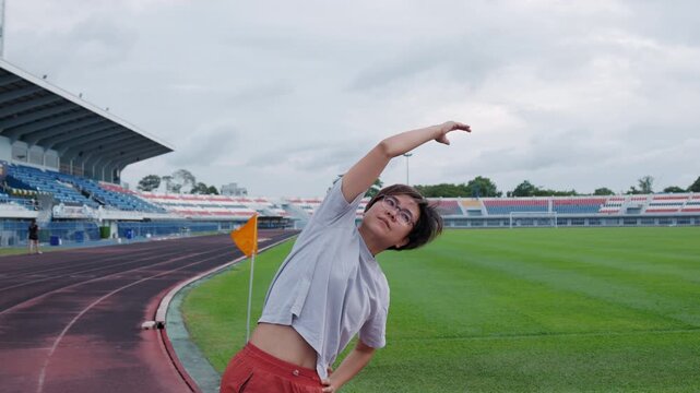 Authentic shot of an active Asian woman in sportswear stretching her arms on a running track. An empty stadium, green soccer field, and waving yellow flag provide excellent copy space.
