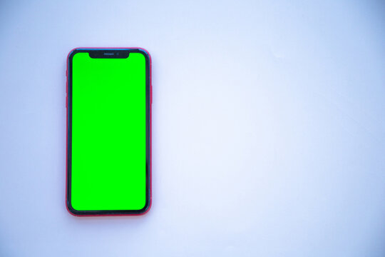 A smartphone with a green screen in a red case lies on a light background on the left. The concept of chroma key, screen replacement, content demonstration, digital technology, modern gadget, mobile