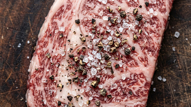 A close-up macro shot of a raw, highly marbled beef steak seasoned with coarse sea salt and cracked black peppercorns on a dark wooden cutting board.
