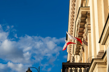 Close-up of the facade of a historic beige tenement building with decorative architectural details, featuring two Polish flags waving against a deep blue sky with white clouds on a sunny day. © Adam