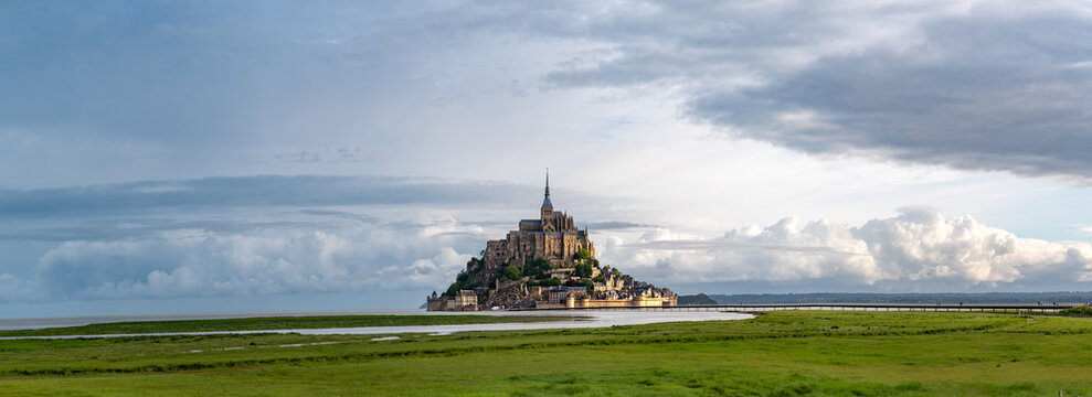 Road to Mont-Saint-Michel during high tide with rising water, tidal phenomenon on Atlantic's ocean coast, view on UNESCO heritage site and iconic Normandy landmark and green pastures
